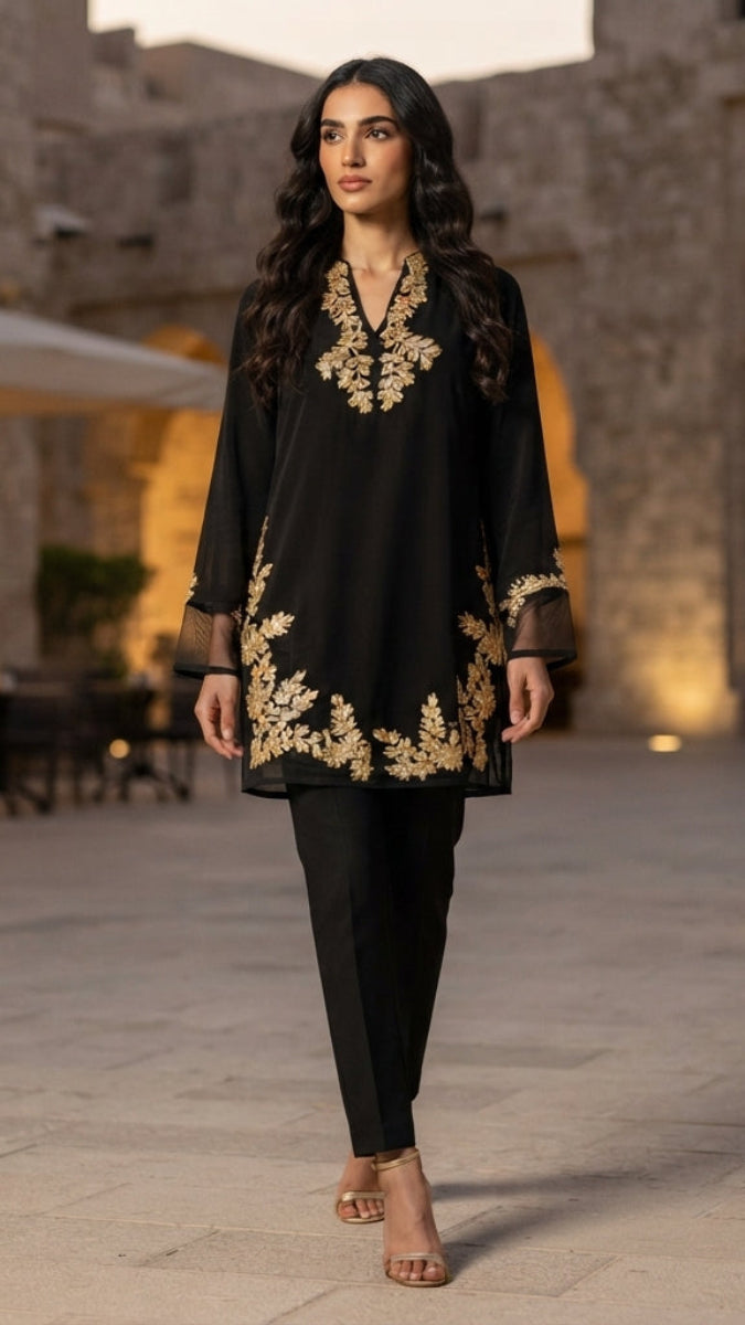 A woman models a black kurta with gold floral embroidery and matching black trousers in an outdoor, stone-paved street setting at dusk.