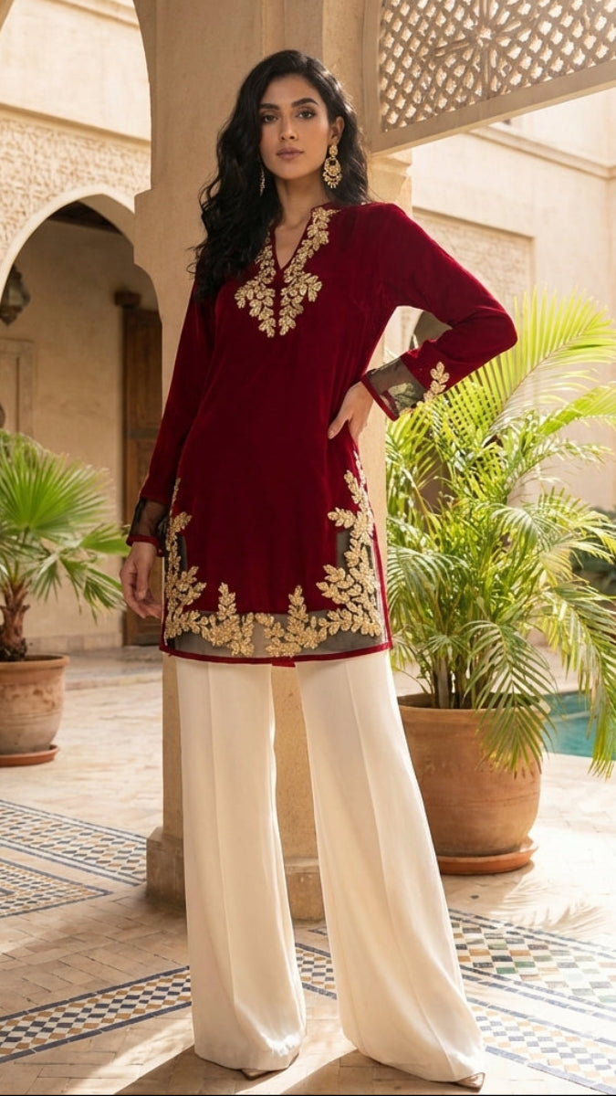 Woman in a maroon embroidered top and white pants standing in a courtyard with plants and a pool.