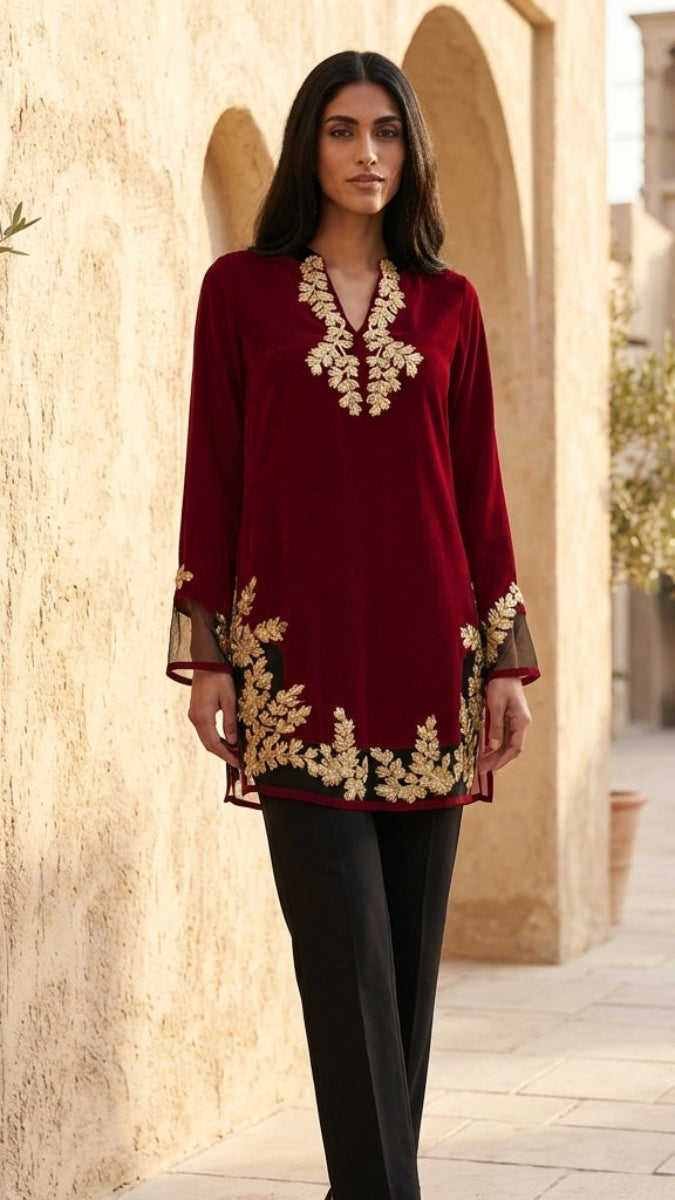 Woman wearing a red embroidered top and black pants standing in an outdoor setting with beige walls and plants.