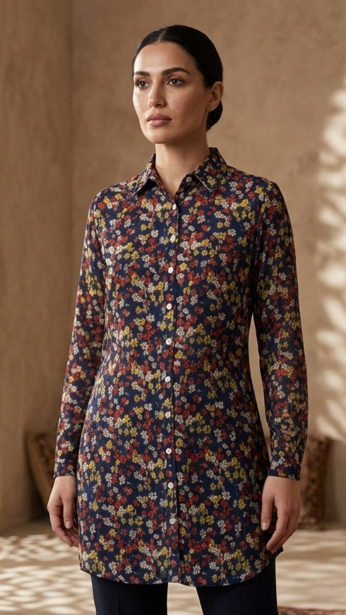 A woman stands indoors wearing a long-sleeve button-front tunic top with an all-over dense floral print in navy, red, yellow, and cream tones, paired with dark pants. The setting has warm beige walls, decorative lanterns, and shadow patterns cast across the floor from a screen.