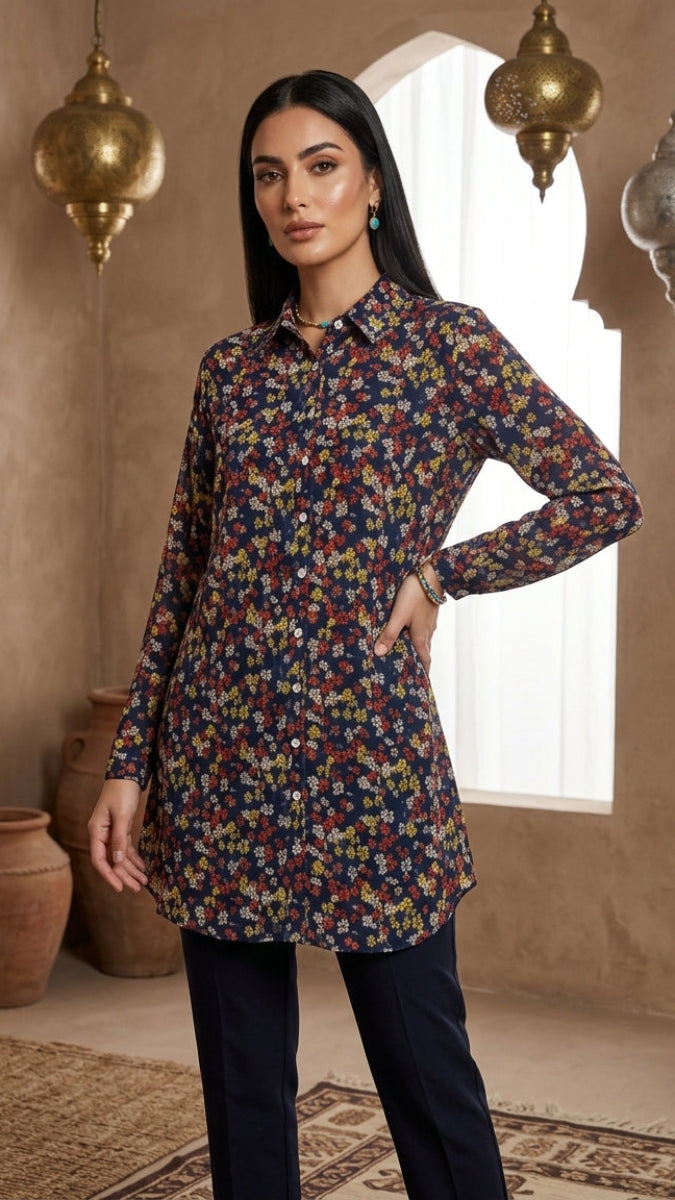 A woman models a long-sleeve button-up tunic top with a dense multicolor floral print on a dark navy base, paired with dark navy trousers, set against a warm, rustic interior with hanging lanterns.