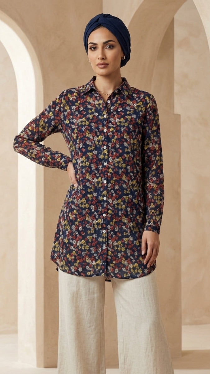 A woman wearing a long-sleeve button-up tunic top with a dense multicolor floral print on a dark navy base, paired with light beige wide-leg trousers and a navy blue headscarf, standing in a beige architectural setting with arches.