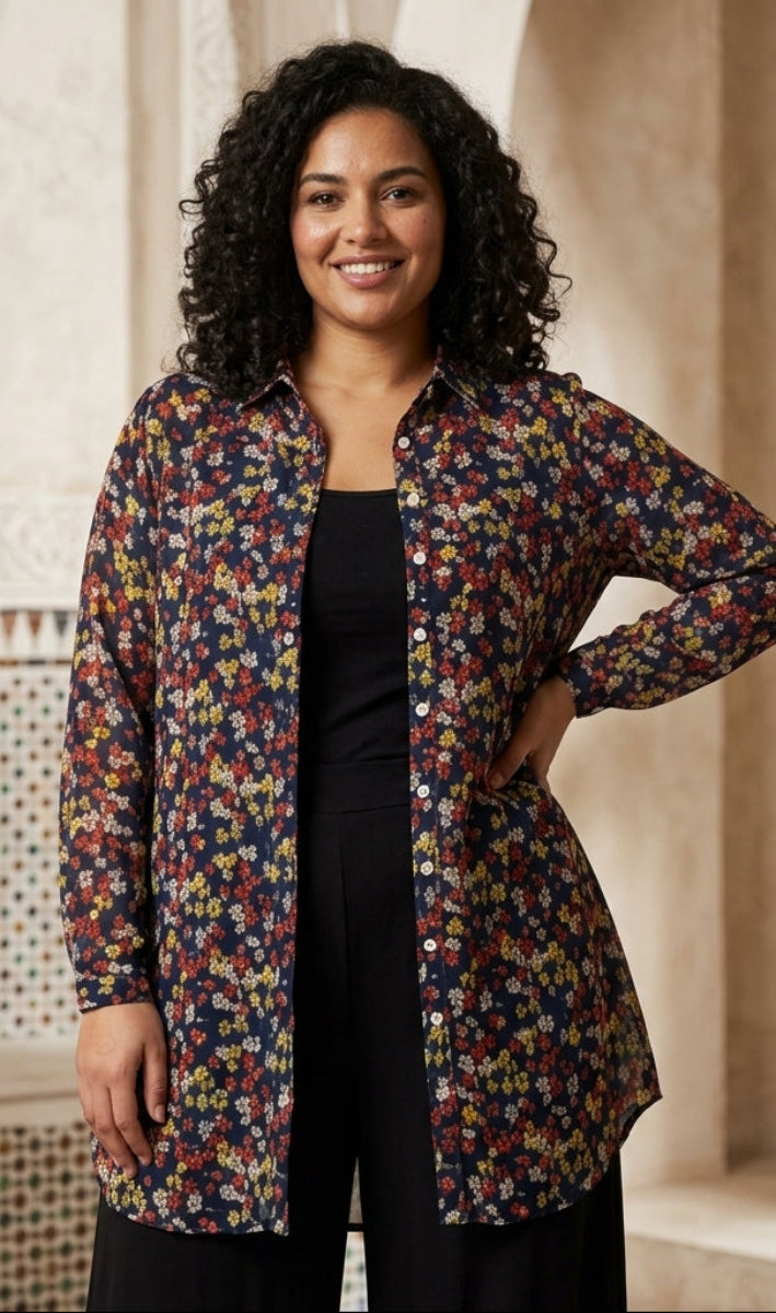A woman with curly black hair stands indoors wearing a long-sleeve button-front shirt with a dense multicolor floral print over a dark navy base, layered over a black top and black pants. The setting features warm beige stone arches and carved wooden decor.