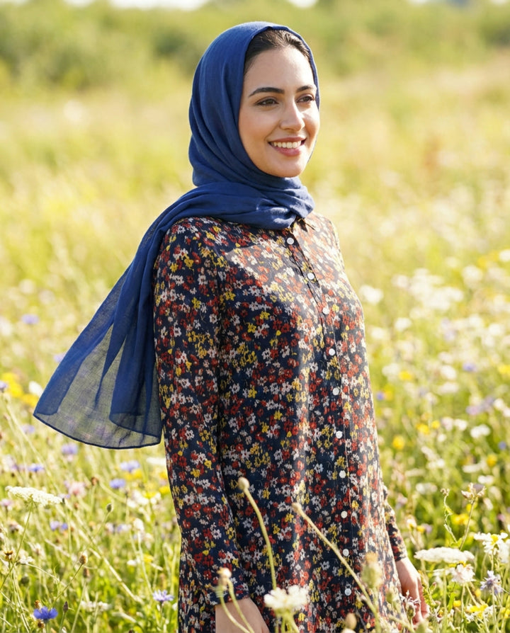 Woman wearing a blue hijab and floral dress standing in a field of wildflowers.