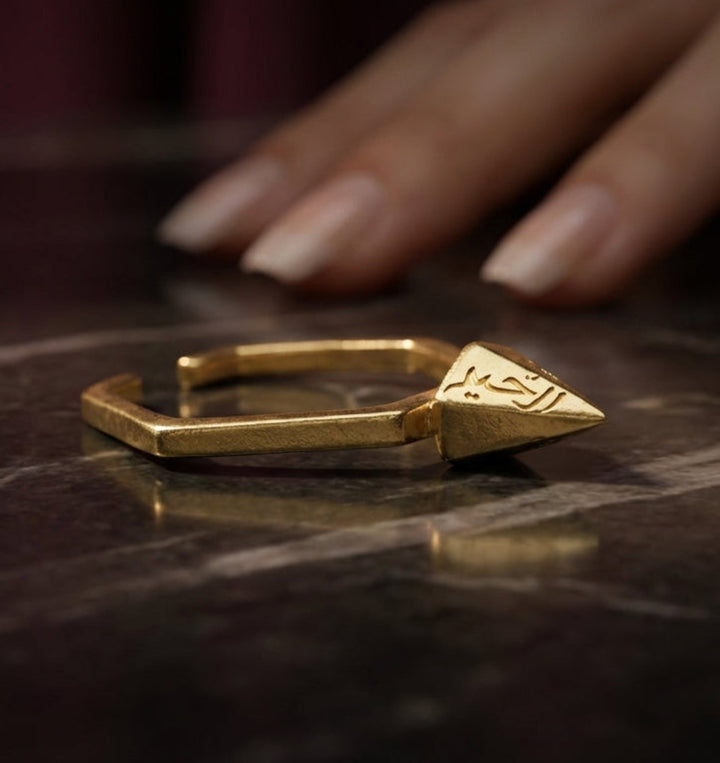Gold ring with geometric design on a dark marble surface, hand partially visible.