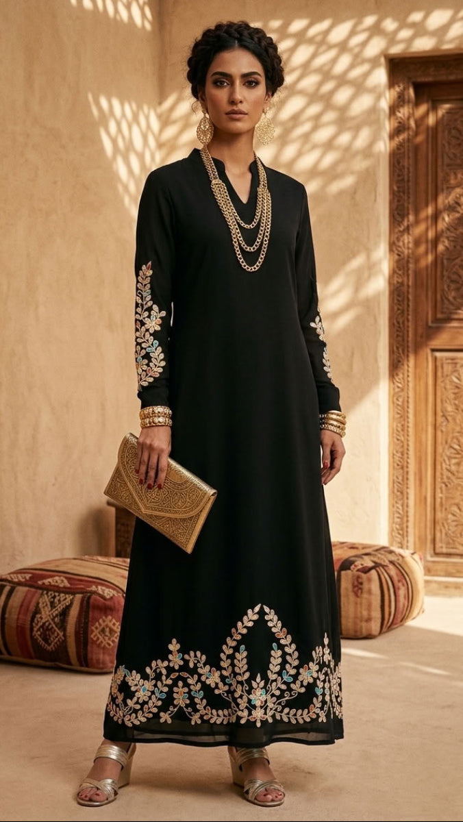 A woman models a black long kurta with intricate gold and beige floral embroidery on the sleeves and hem, styled with gold jewelry and a gold clutch in a warm, sunlit courtyard setting.