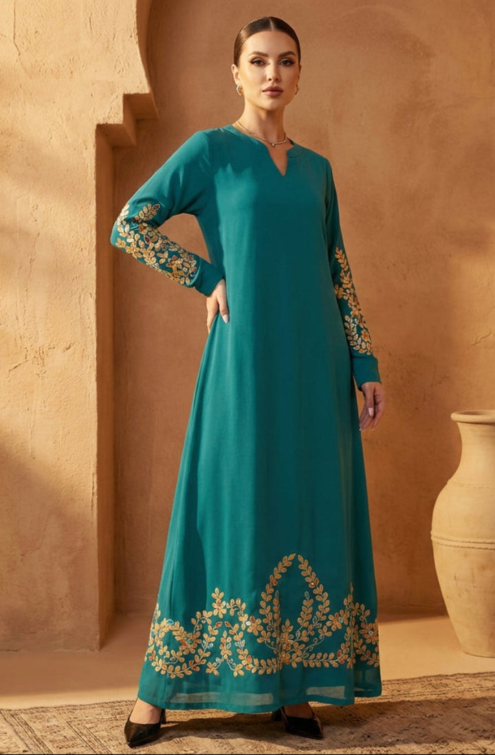 Woman in a teal dress with gold embroidery standing against a beige wall.