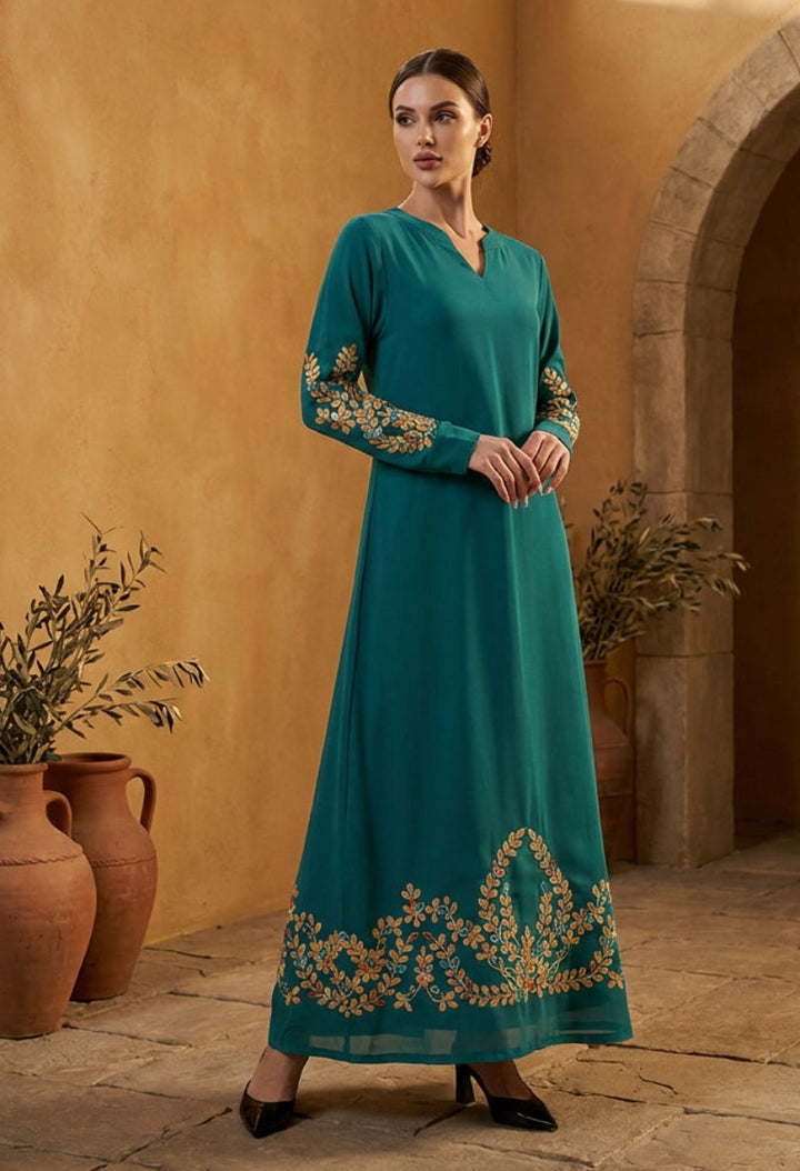 Woman in a teal dress with gold embroidery standing in a sunlit room with stone walls and potted plants.