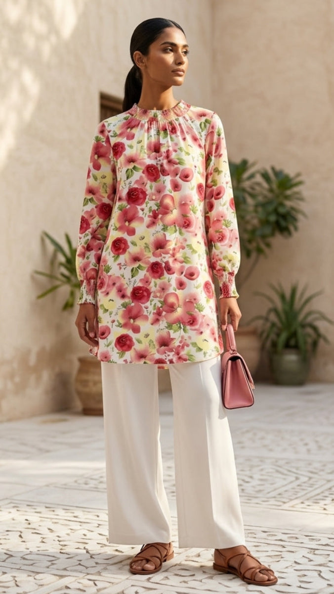 Woman in a floral top and white pants standing in an outdoor setting with a building and plants in the background.