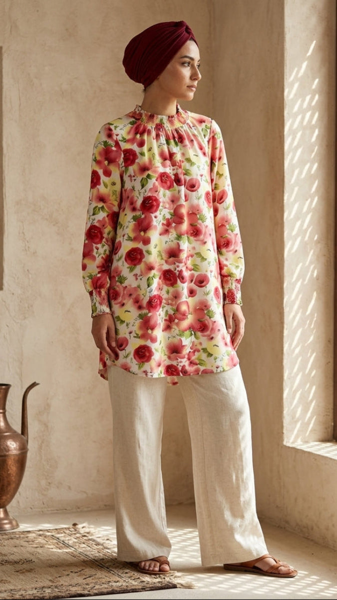 Woman wearing a floral top and white pants standing in a sunlit room with a textured wall and window.