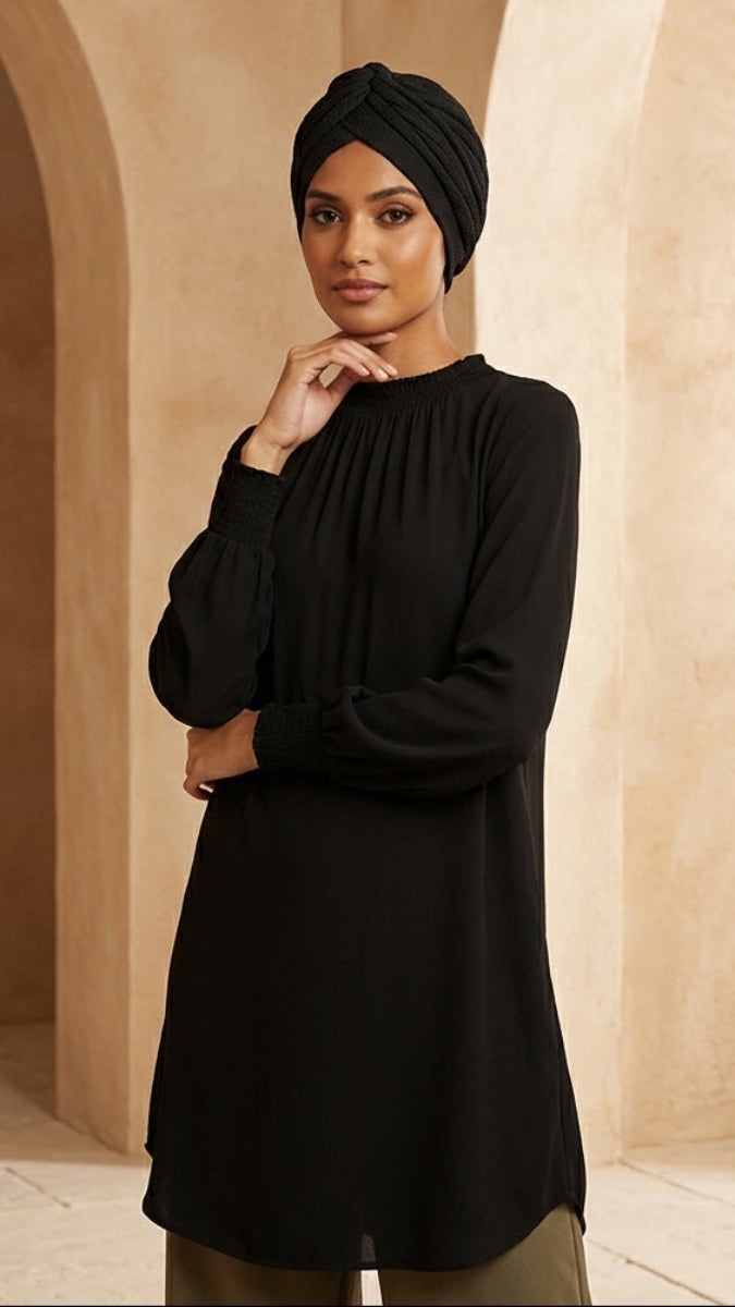 Woman wearing a black long-sleeve top and headscarf in a warm-toned architectural setting.
