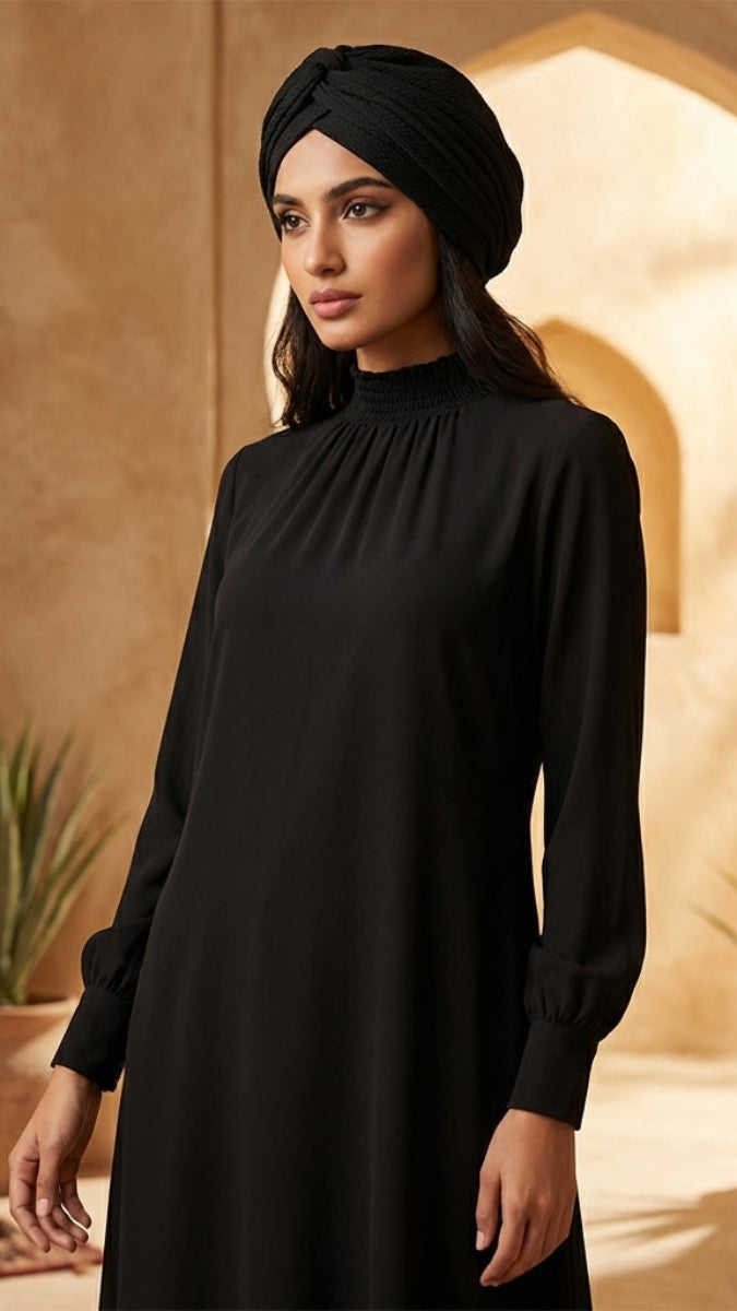 Woman in a black dress and headscarf standing in a warm-toned interior setting with plants.