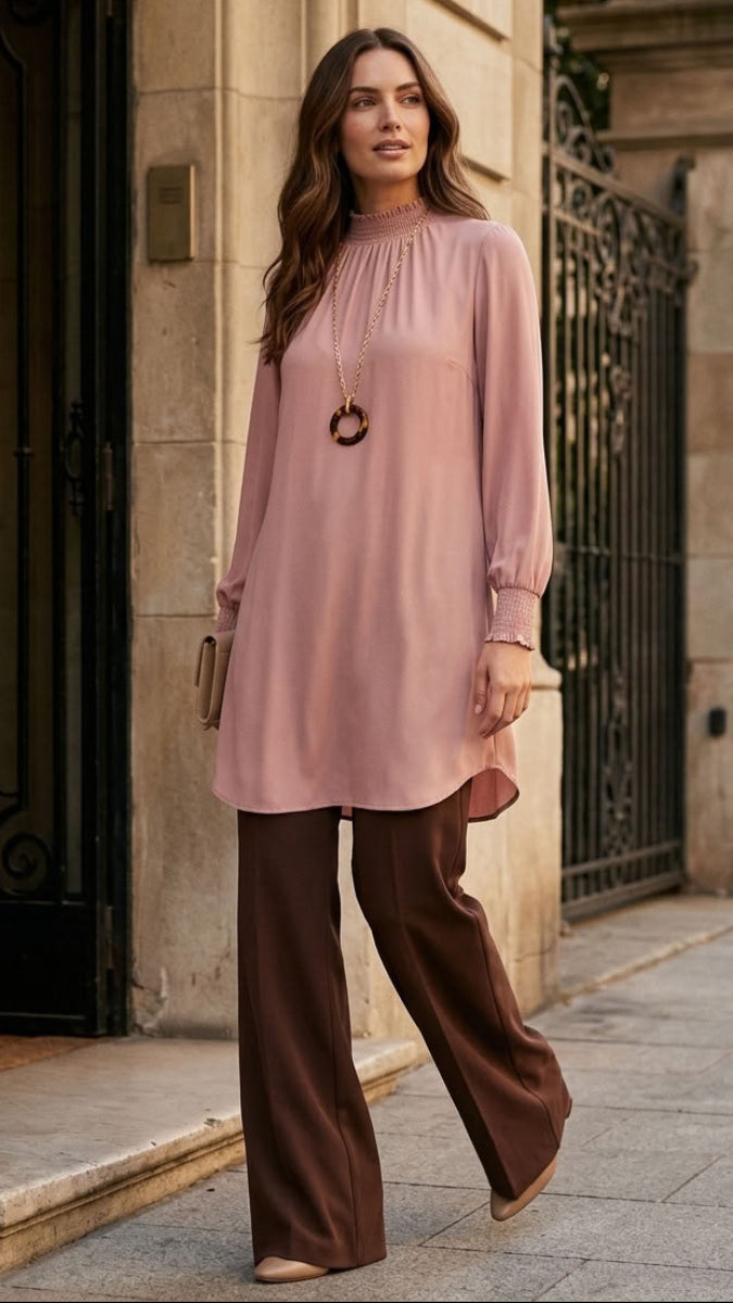 A woman stands on a city sidewalk wearing a dusty pink long-sleeve tunic top with a gathered neckline and brown wide-leg trousers, posed in front of a stone building with black gates.
