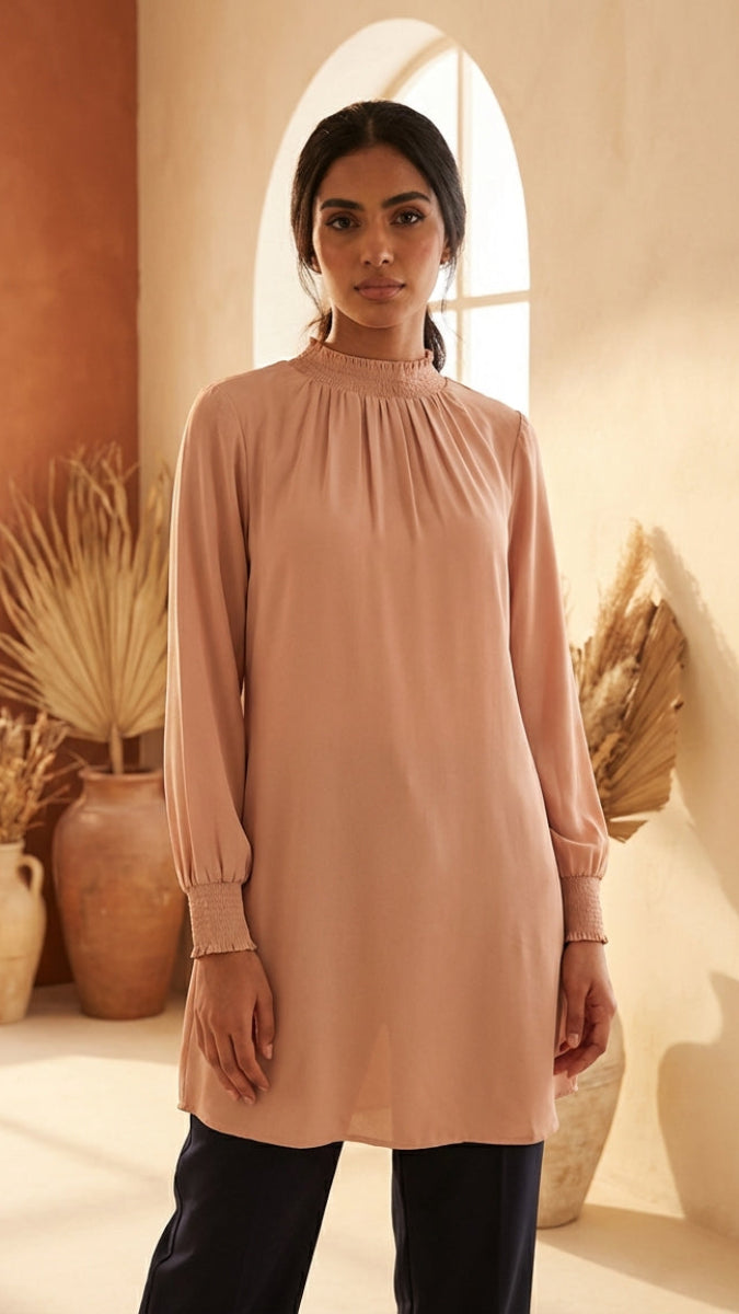 Woman wearing a long-sleeve, high-neck beige dress in a warm-toned room with arched windows and decorative plants.