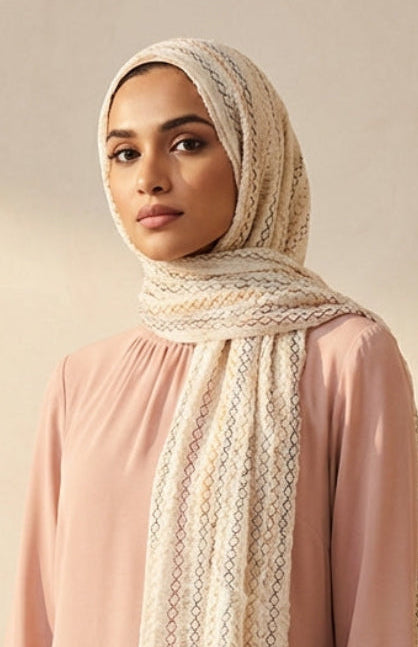 Woman wearing a pink long-sleeve top and a beige hijab with a patterned scarf against a beige wall.