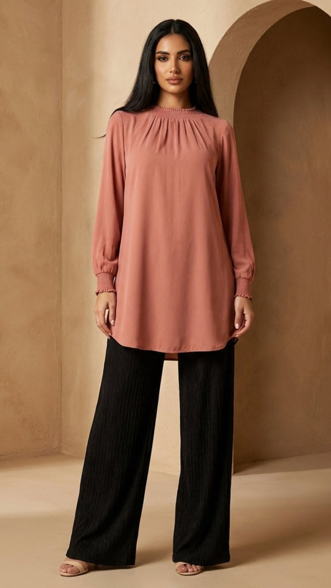 Woman wearing a pink long-sleeve top and black pants in a beige interior setting