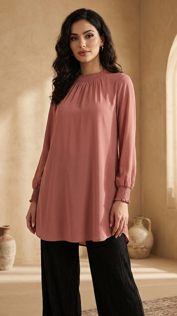 Woman wearing a pink long-sleeve top and black pants in a warm-toned room with vases.