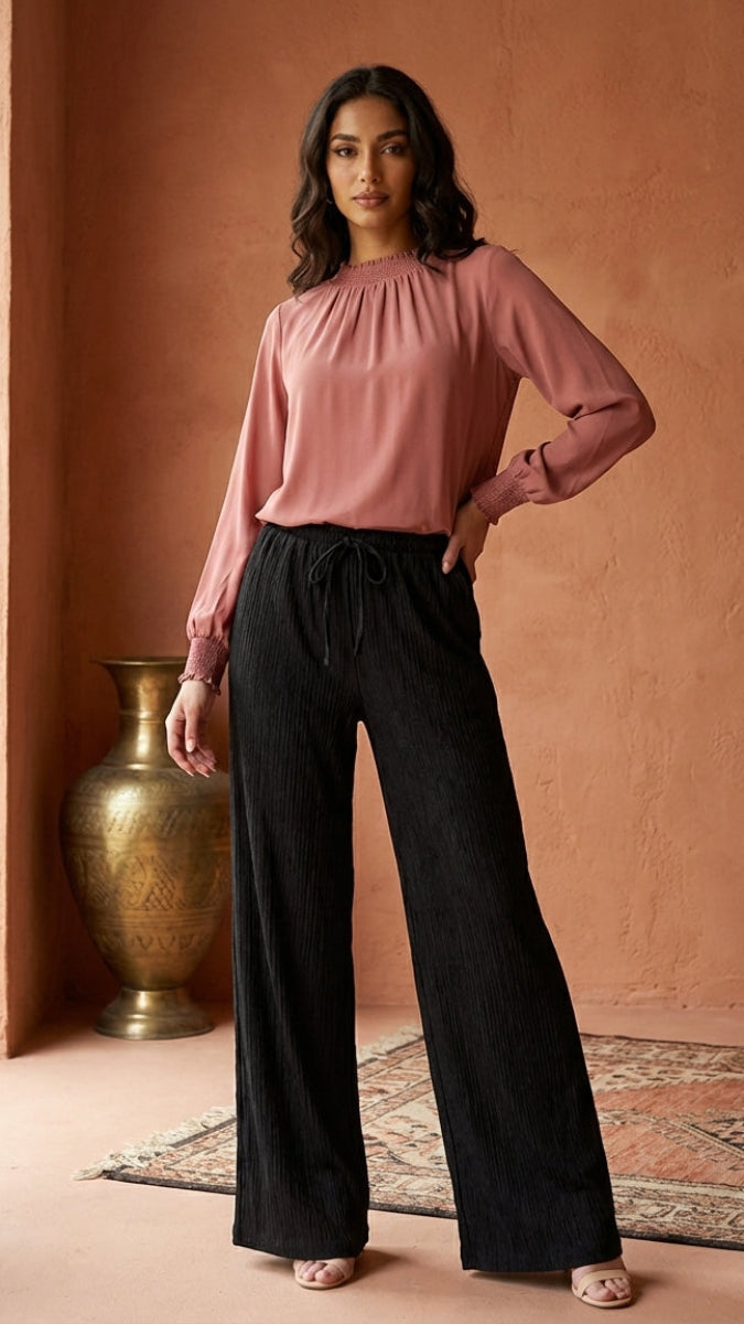 Woman wearing a pink blouse and black pants standing against a warm-toned wall.