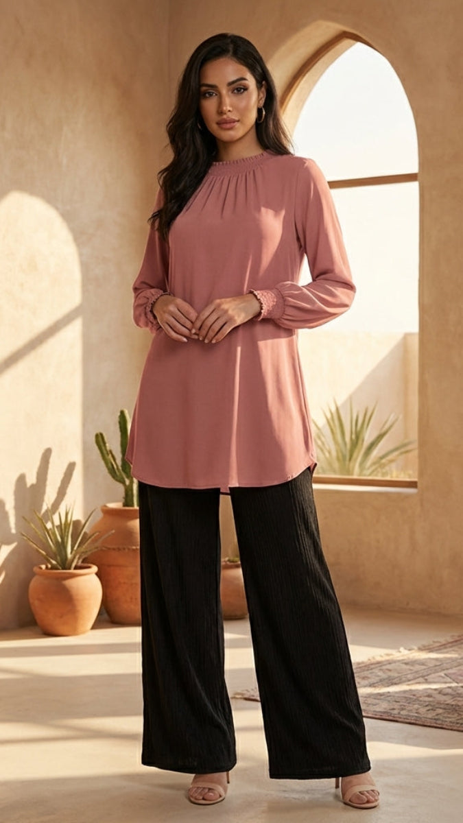 Woman in a pink top and black pants standing in a sunlit room with arches and plants.
