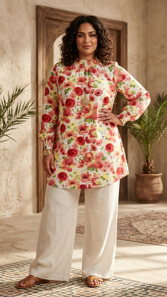 Woman wearing a floral top and white pants in a sunlit room with plants.