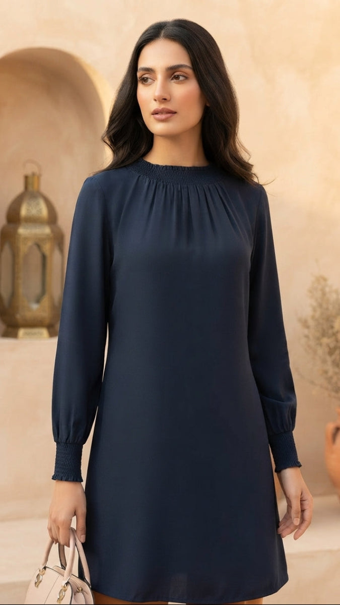 A woman wearing a navy blue long-sleeve shift dress stands in front of a beige stucco wall with an arched niche and a potted plant. She is holding a small light beige handbag.