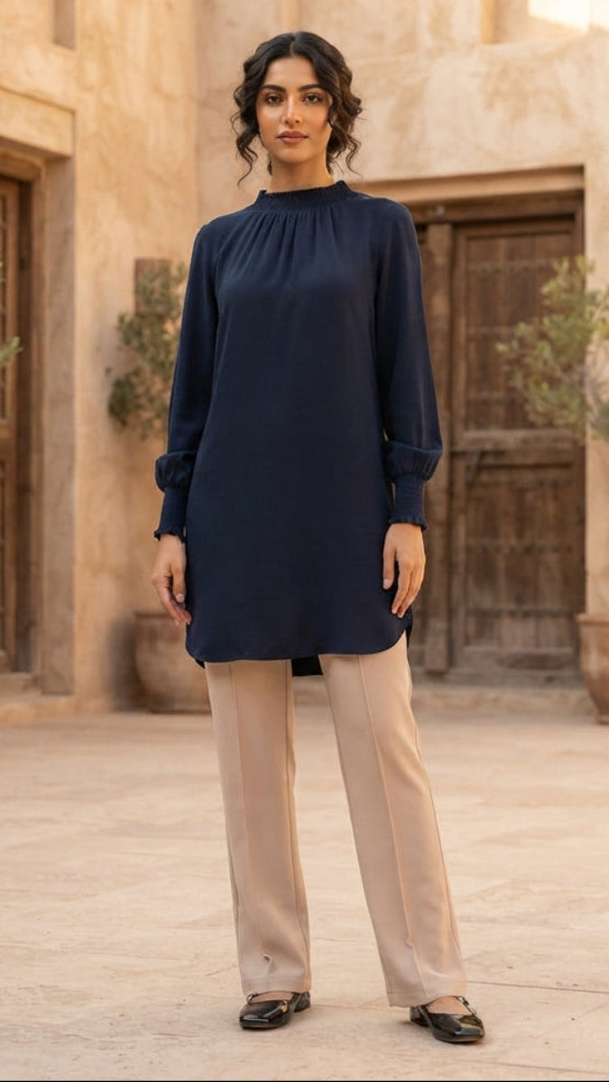A woman stands in a courtyard wearing a navy blue long-sleeve tunic with a gathered neckline and beige wide-leg trousers. The background features warm beige stucco walls, wooden doors, and potted plants.