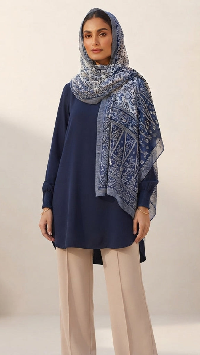 A woman modeling a navy blue long-sleeve tunic with a matching navy and beige patterned hijab, paired with beige wide-leg trousers, set against a warm beige architectural background.