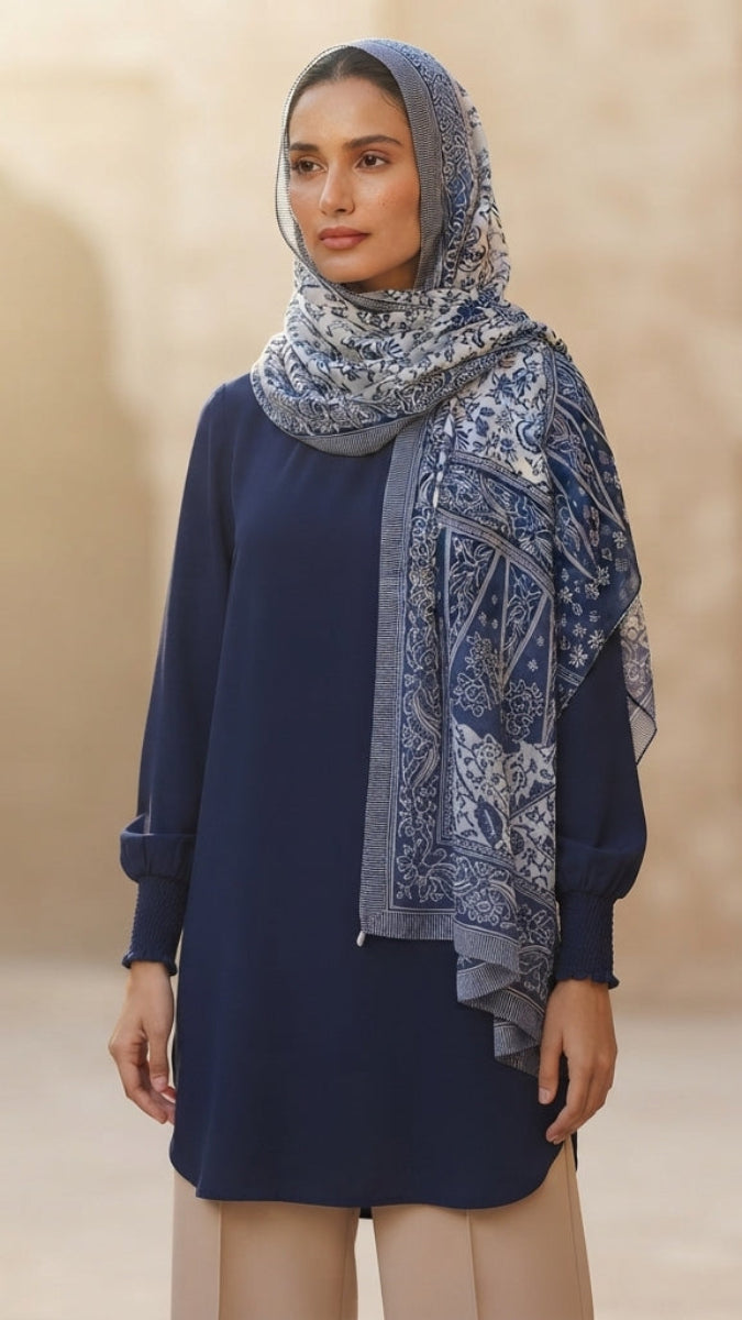 A woman wearing a navy blue long-sleeve tunic and a blue and white patterned hijab, standing in a warm-toned architectural setting.