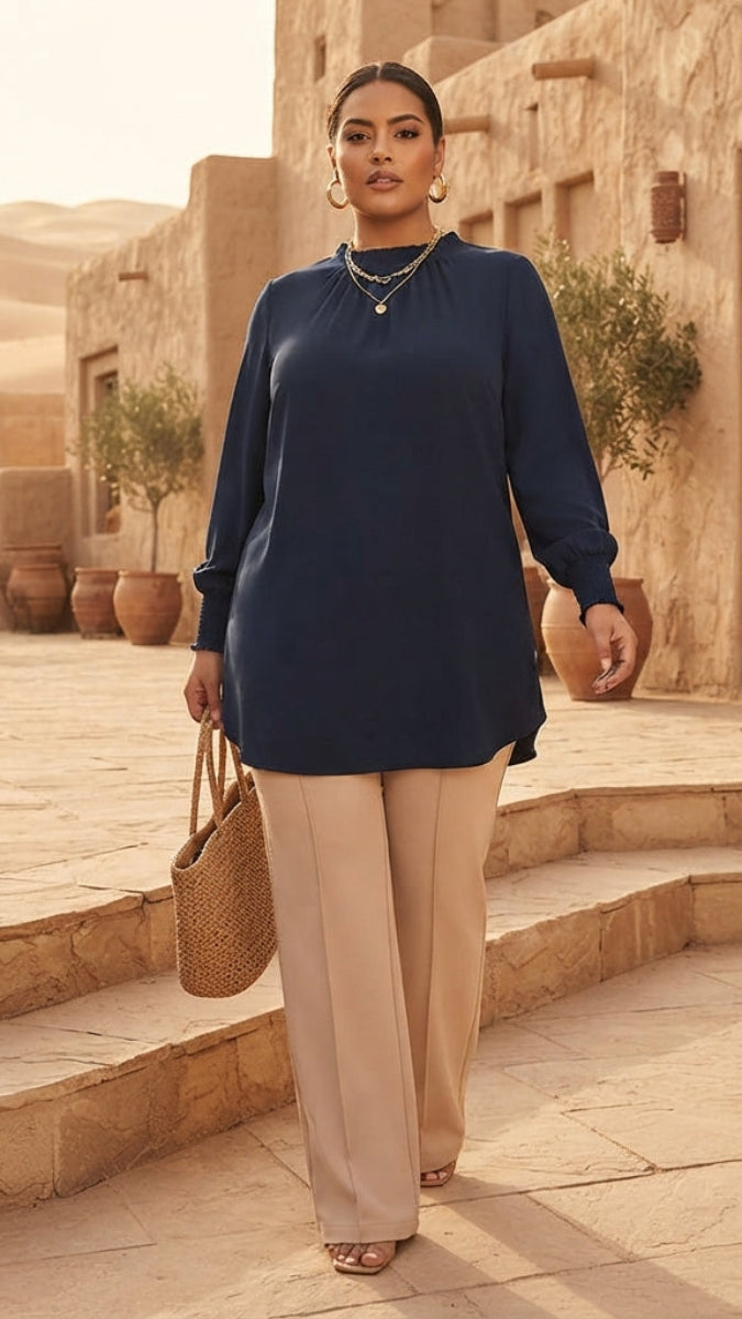 A woman walks down stone steps in a desert village setting, wearing a navy blue long-sleeve tunic top and beige wide-leg trousers, carrying a woven straw handbag.