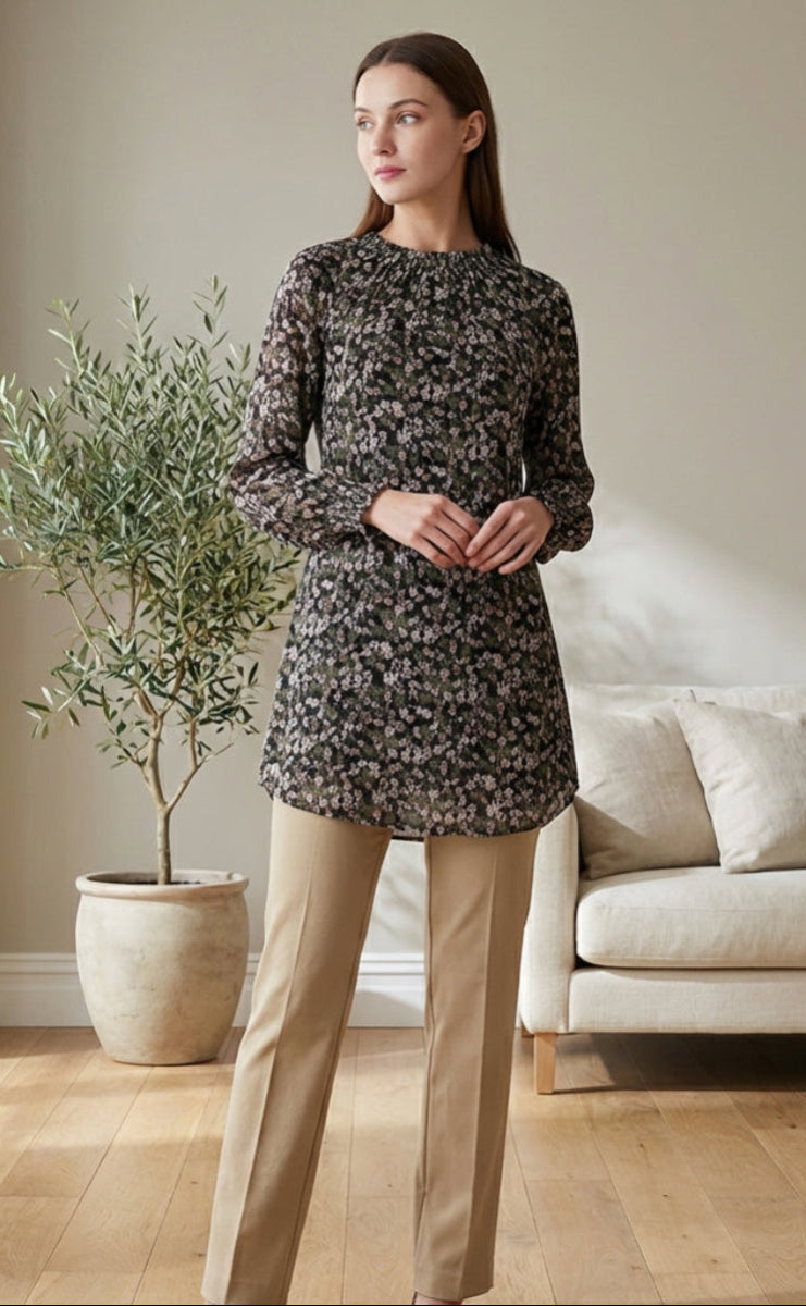 Woman in a floral dress and beige pants standing in a living room with a plant and sofa.