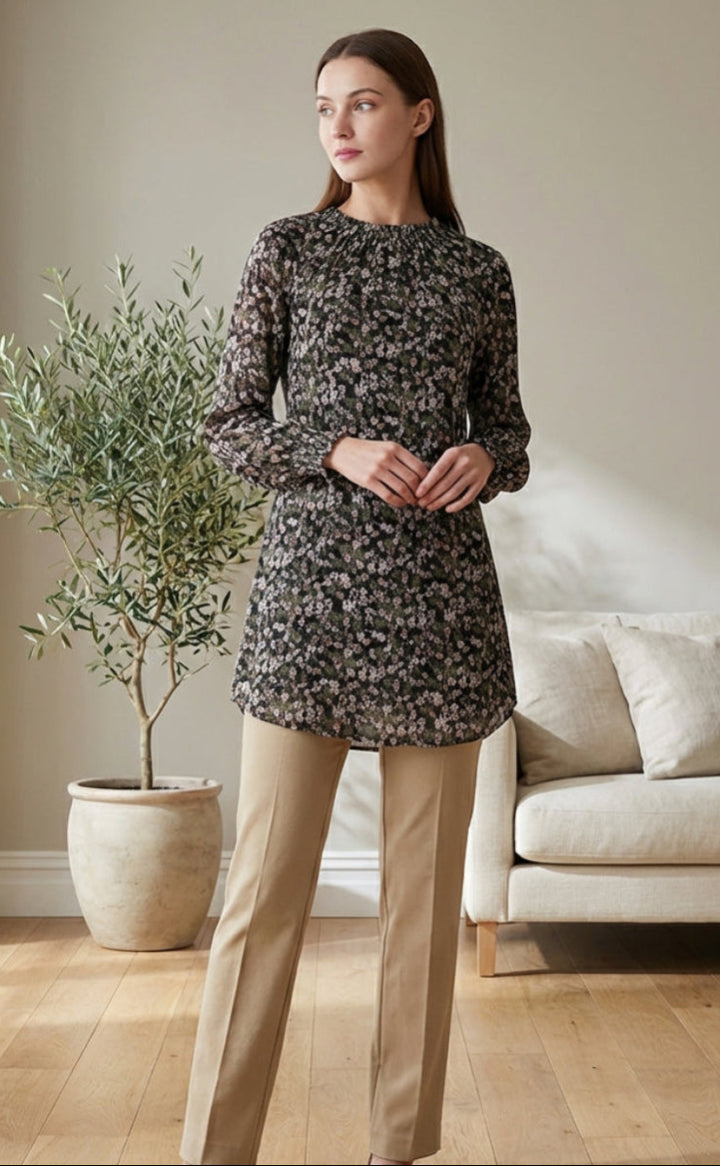 Woman in a floral dress and beige pants standing in a living room with a plant and sofa.