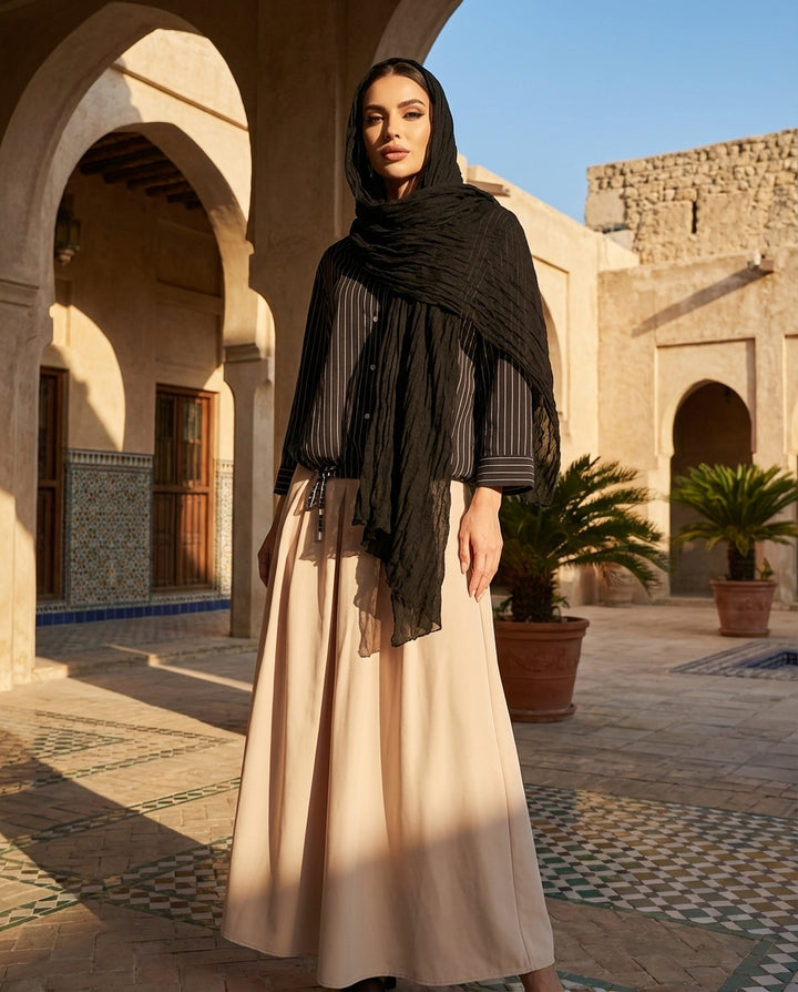 Woman in a hijab and long dress standing in a sunlit courtyard with arches and plants.