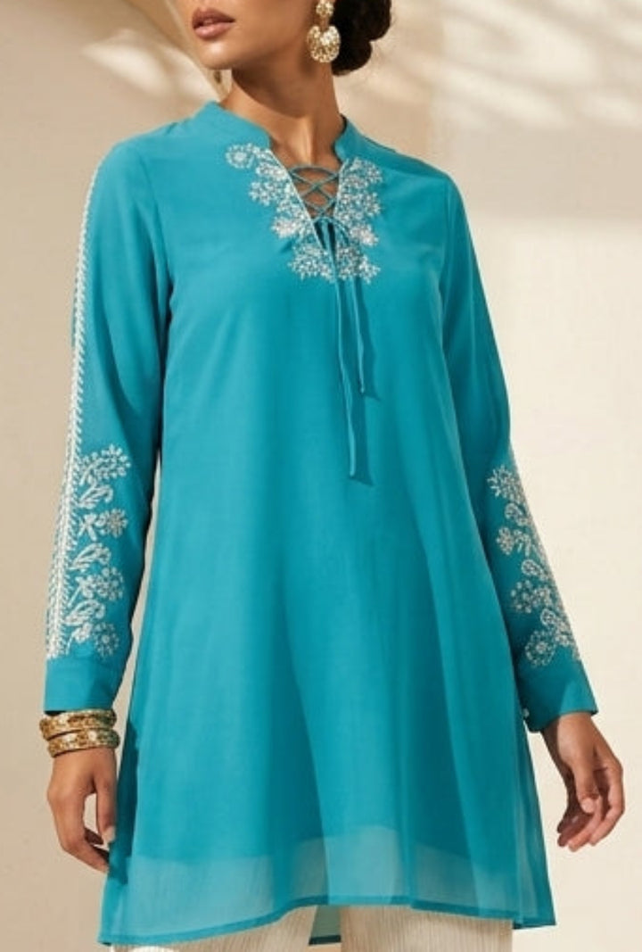 Woman wearing a turquoise embroidered top and white pants in a sunlit room with archway.