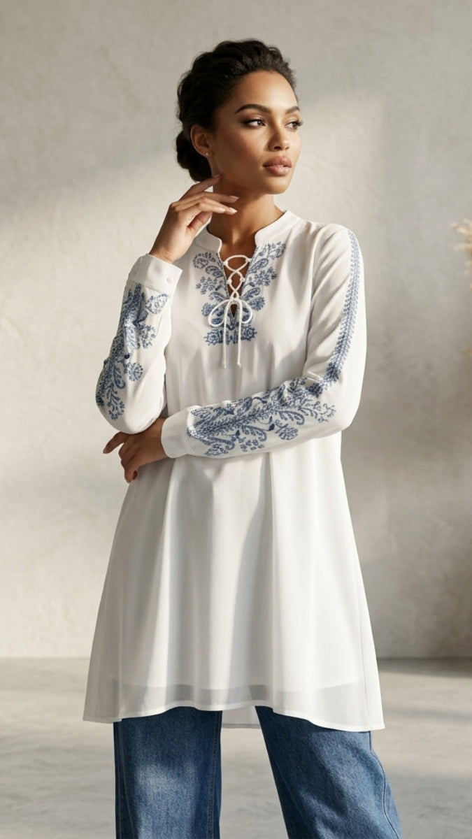 Woman wearing a white embroidered long-sleeve top with blue patterns, standing in a room with a vase of dried plants.
