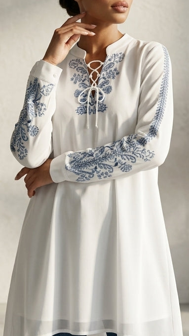 Woman wearing a white embroidered long-sleeve top with blue patterns, standing in a room with a vase of dried plants.