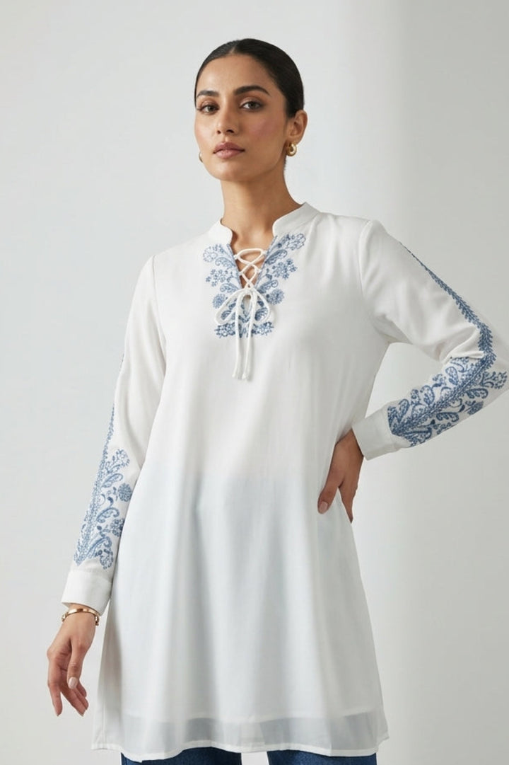 Woman wearing a white embroidered long-sleeve top with blue patterns on a plain background