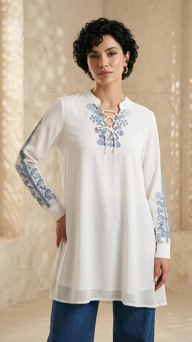 Woman wearing a white embroidered long-sleeve top with blue patterns in a decorative setting.
