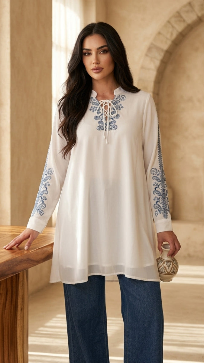 Woman wearing a white embroidered tunic with blue patterns in a sunlit room.