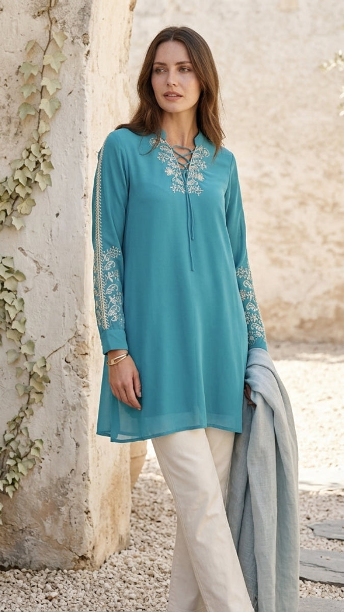 Woman wearing a turquoise embroidered top and white pants standing against a stone wall with greenery.