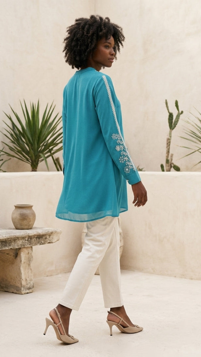 Woman in a turquoise top and white pants walking outdoors with plants and a bench in the background.