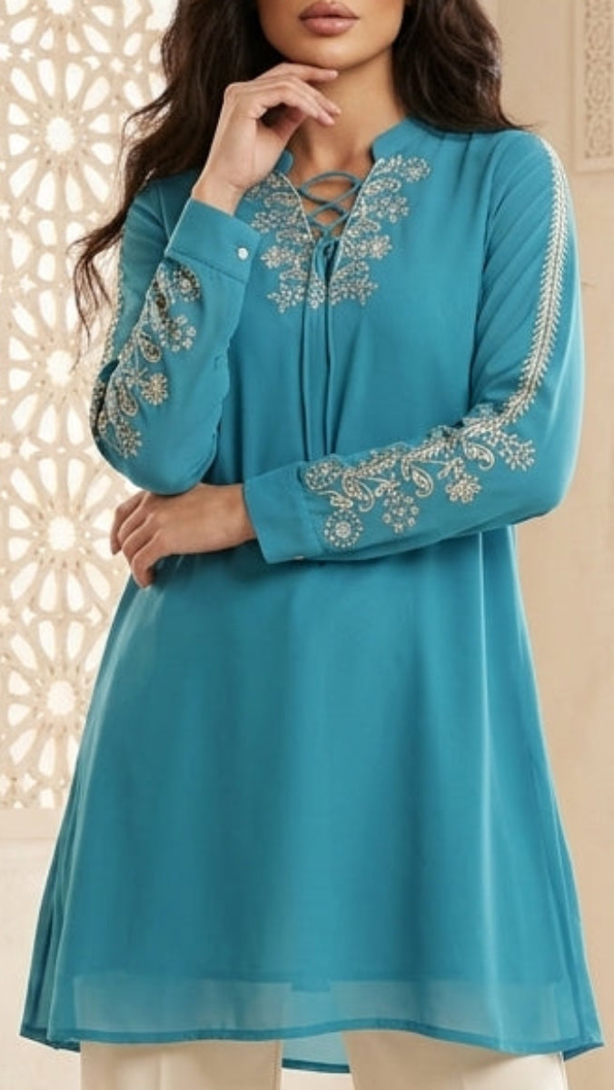 Woman wearing a blue embroidered top and white pants in a decorative room with architectural elements.