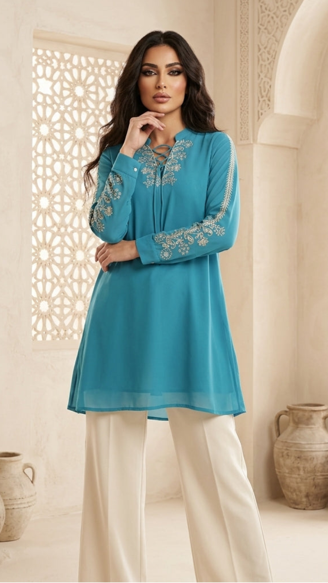 Woman wearing a blue embroidered top with white pants in an ornate room.