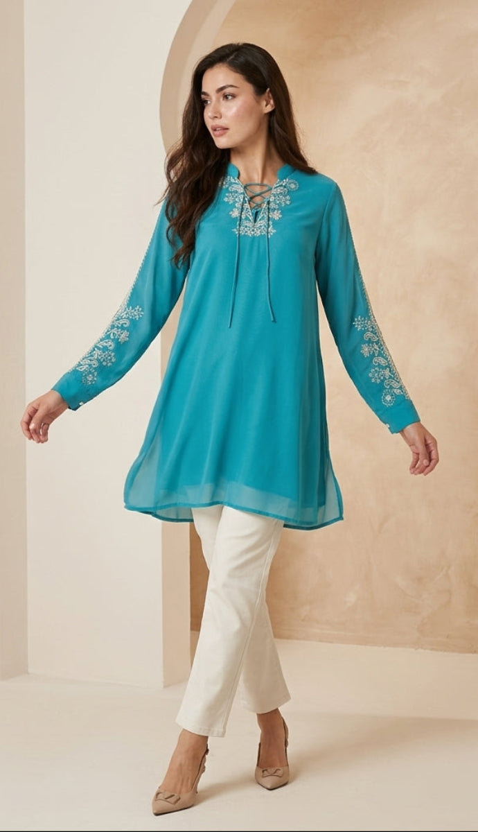 Woman wearing a blue embroidered top and white pants in a neutral setting