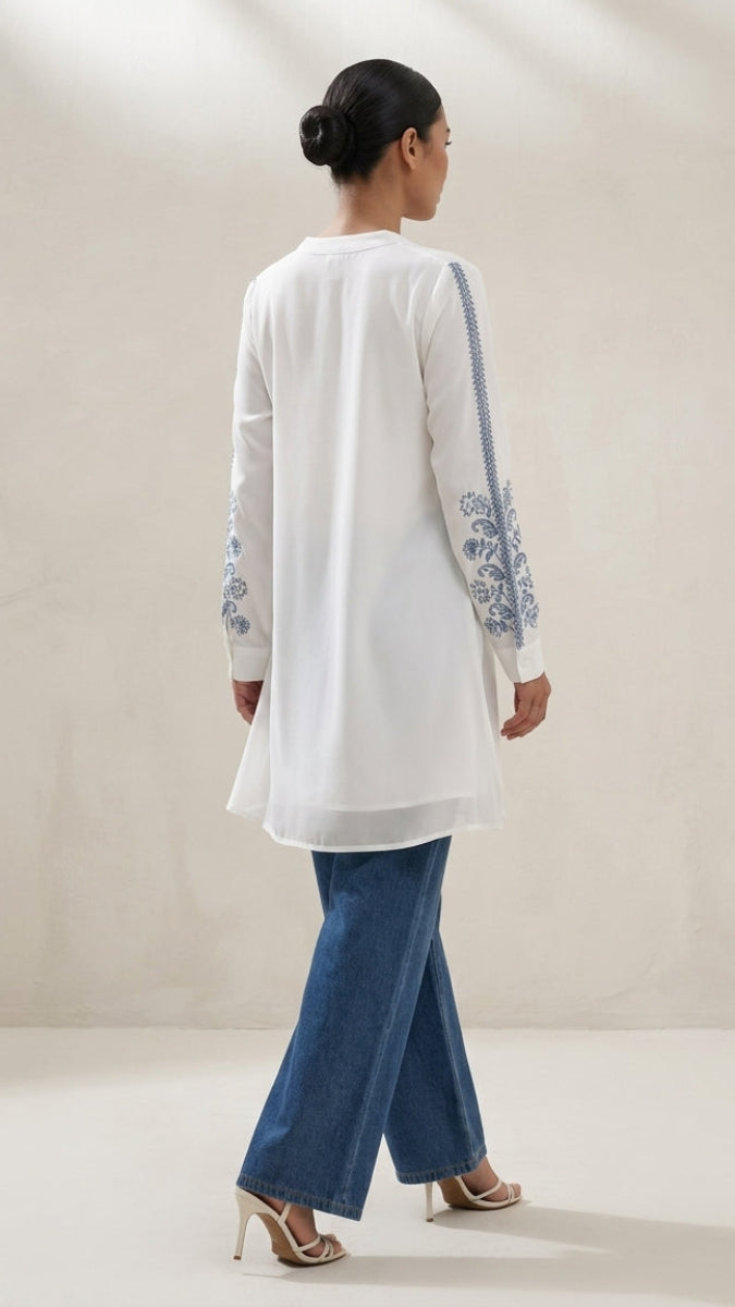 Woman wearing a white embroidered top and blue jeans against a plain background