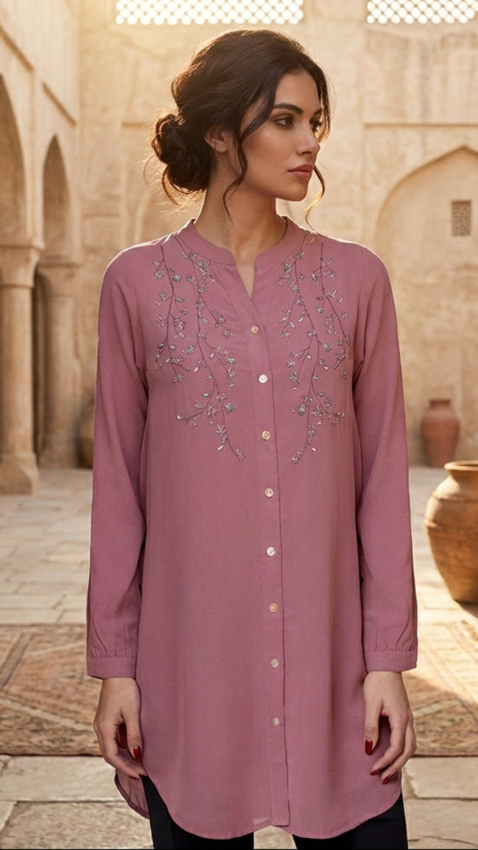 Woman wearing a pink embroidered dress in a sunlit courtyard with arches and pots.