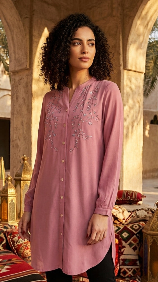 Woman in a pink embroidered dress standing in a courtyard with palm trees and decorative elements.