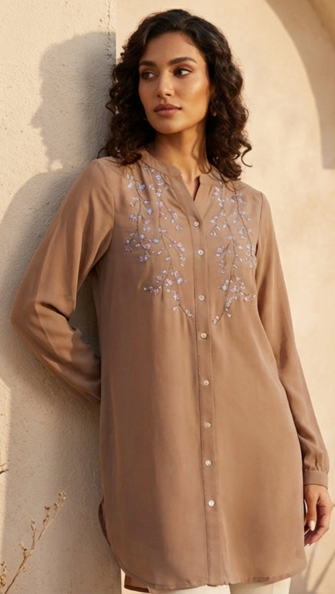 Woman wearing a brown embroidered shirt against a beige wall.