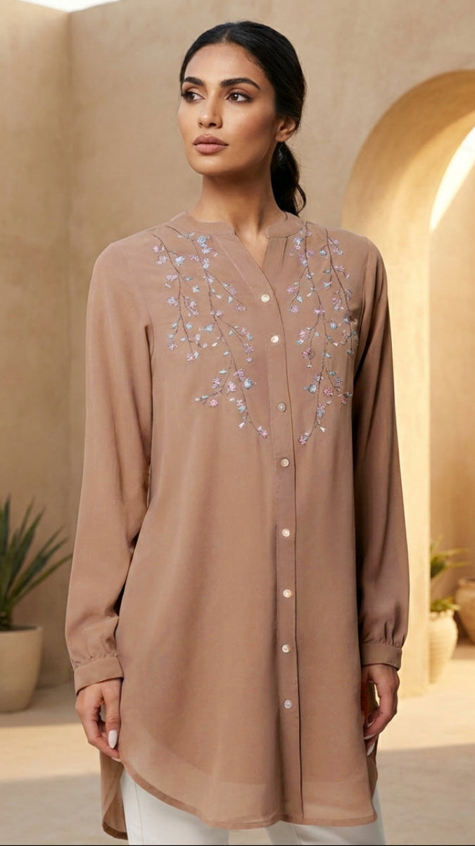 Woman wearing a long, light brown shirt with decorative elements in an indoor setting.