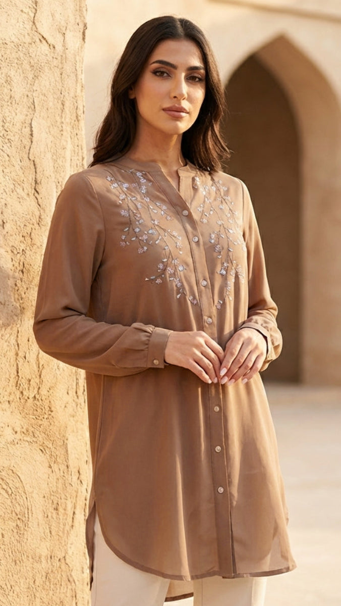 Woman wearing a brown embroidered shirt against a beige wall with an archway.