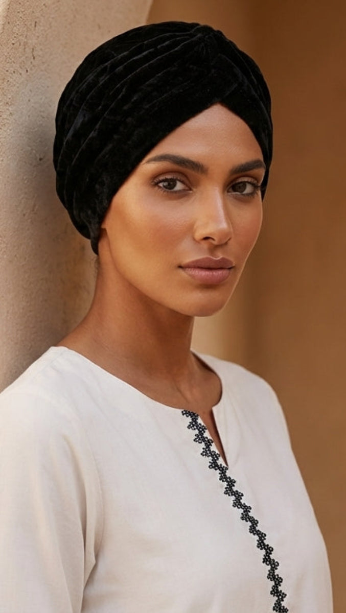 Woman wearing a black turban and white outfit standing against a beige wall with plants in the background